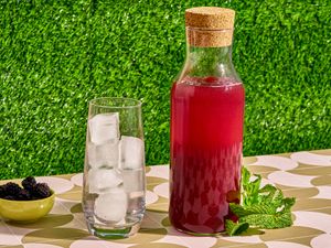 A carafe filled with blackberry liqueur next to a glass of ice, a small bowl of blackberries, and fresh mint sprigs, all on a patterned tablecloth