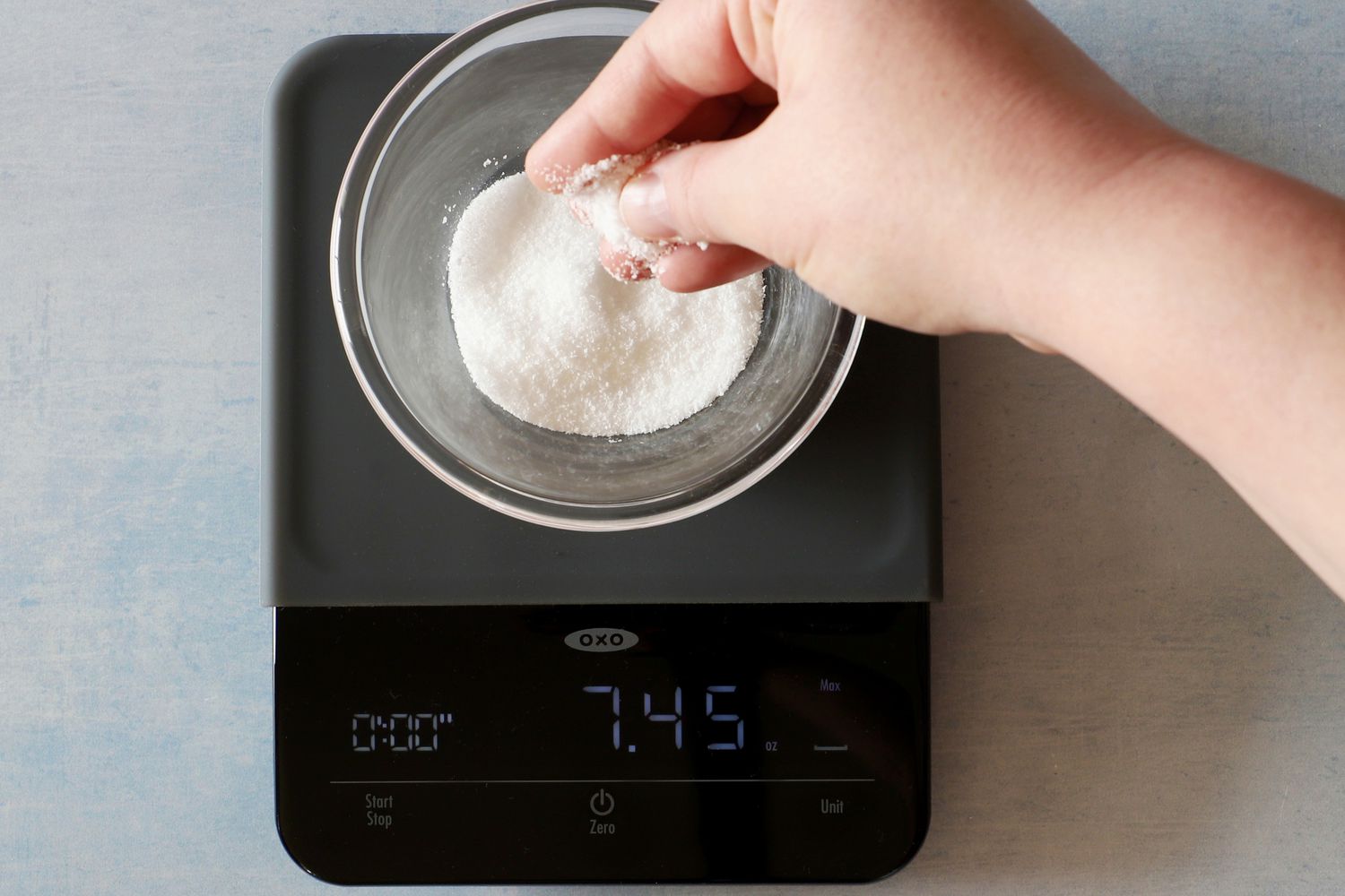 A person measuring sugar into a small glass mixing bowl set on a kitchen scale