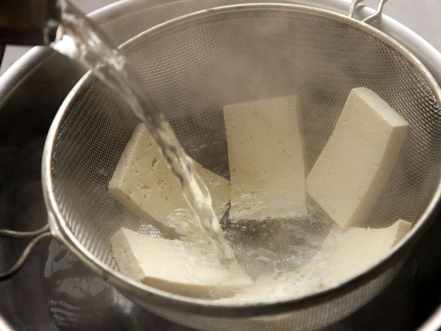 Slabs of tofu are arranged in a fine-mesh strainer and showered with boiling water.