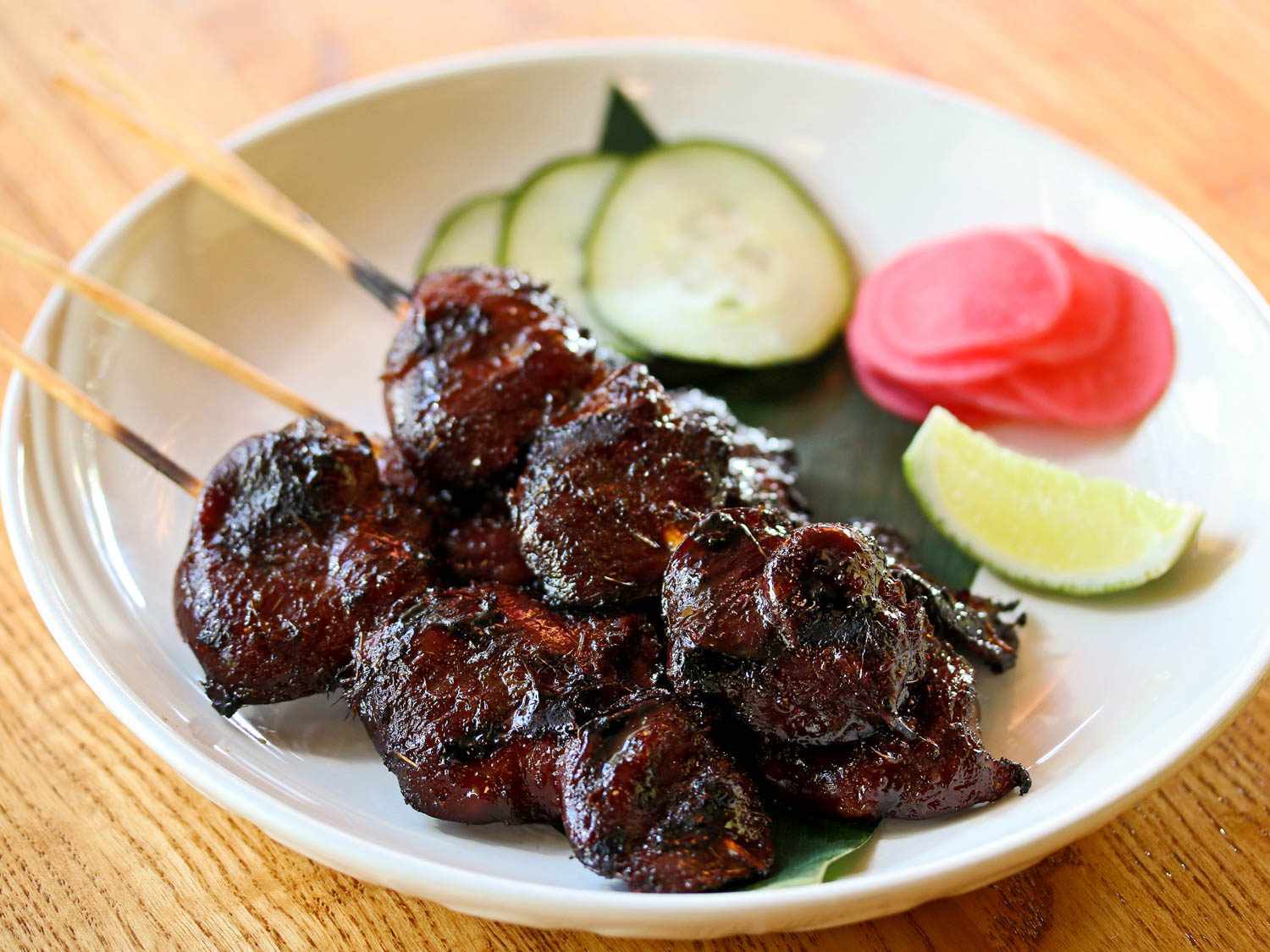 Dark grilled chicken hearts (piing hua jai kai) on a wooden skewer, on a plate with lime, cucumber, and other vegetables in the background.