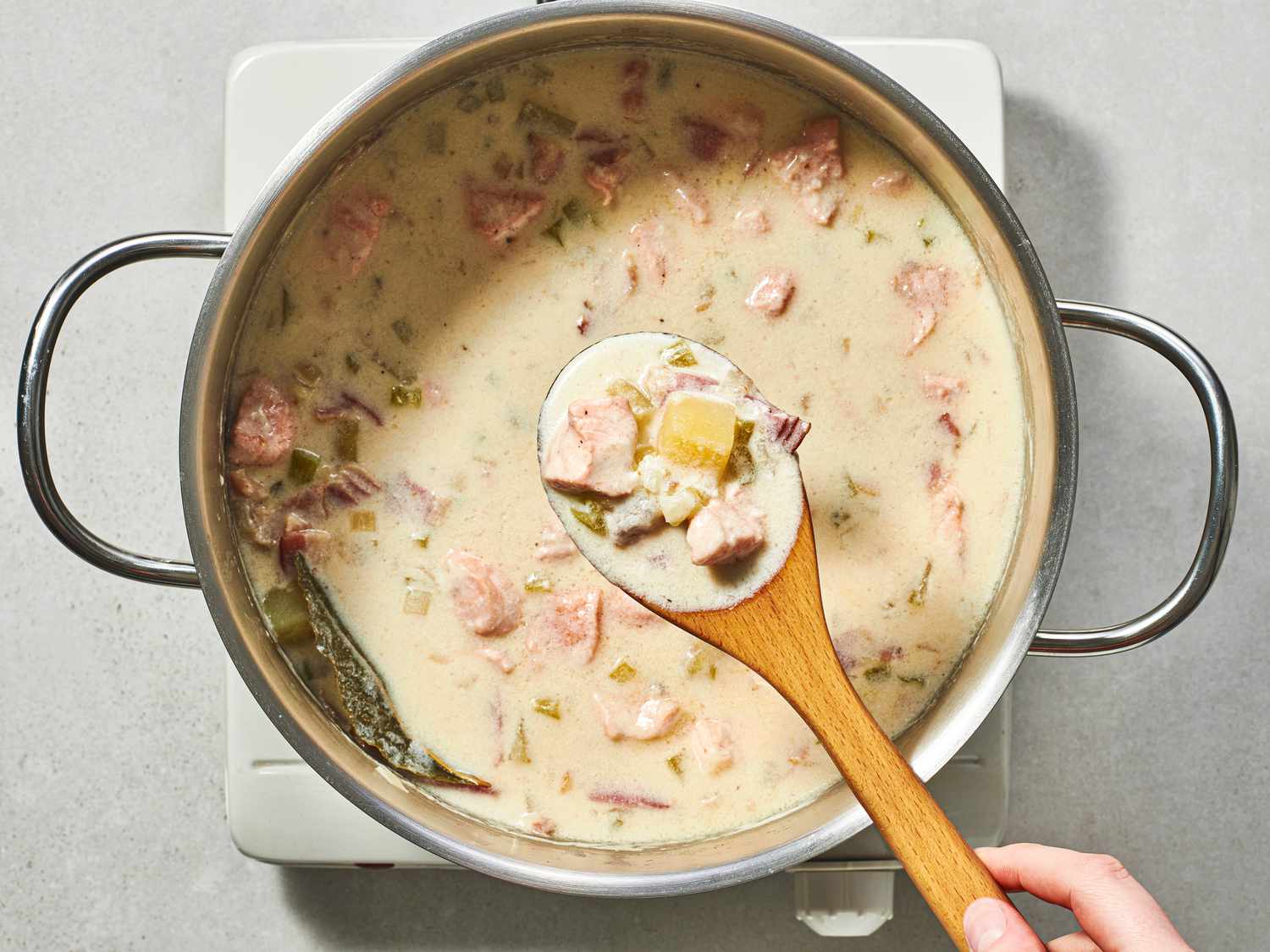 A close-up image of a wooden spoon lifting a spoonful of chowder out of the inside of the stainless steel pot.
