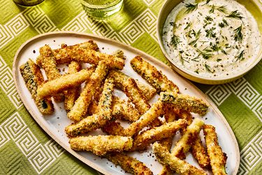 Baked Zucchini fries on a white dish, with a bowl of dip on the side. The table has green printed table cloth and two glasses of water. 