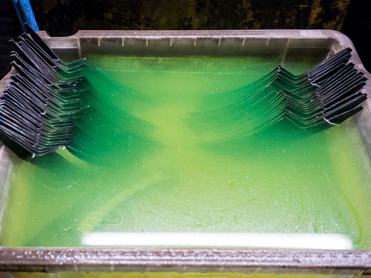 Dozens of finished knife blades are held in a pan full of greenish liquid.