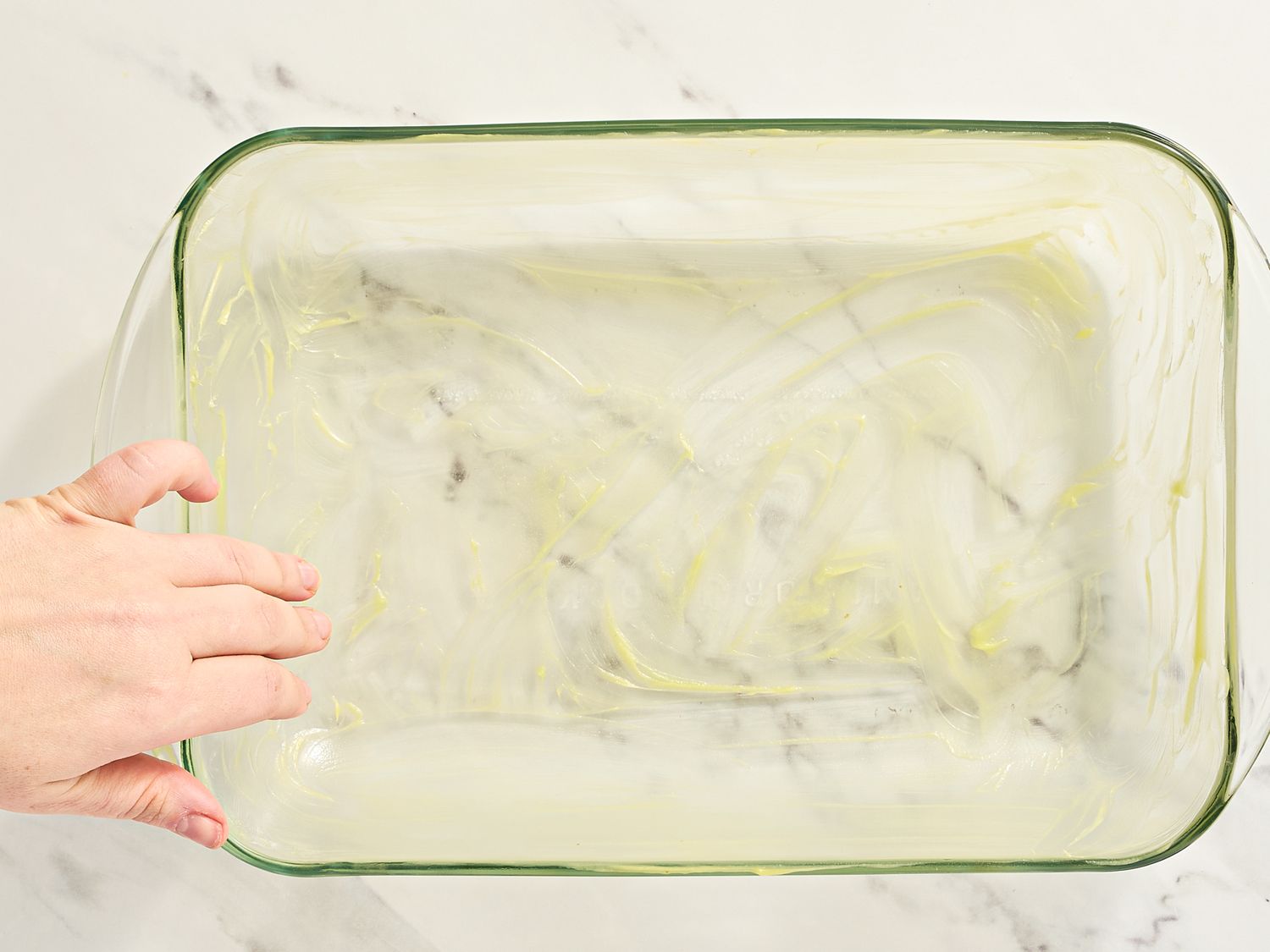 A rectangular glass baking dish being greased by a hand