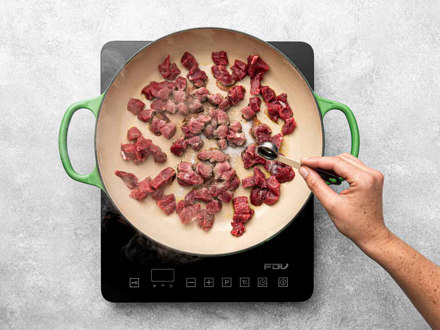 Beef chunks cooking in a pan on an induction stove, a hand stirring with a spoon.