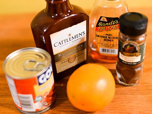 A bottle of barbecue sauce and honey, flanked by a jar of ancho chile powder, a can of chipotles en adobo, and an orange.