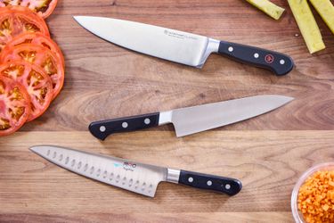 Three chef's knives on a wooden countertop with sliced tomatoes nearby.