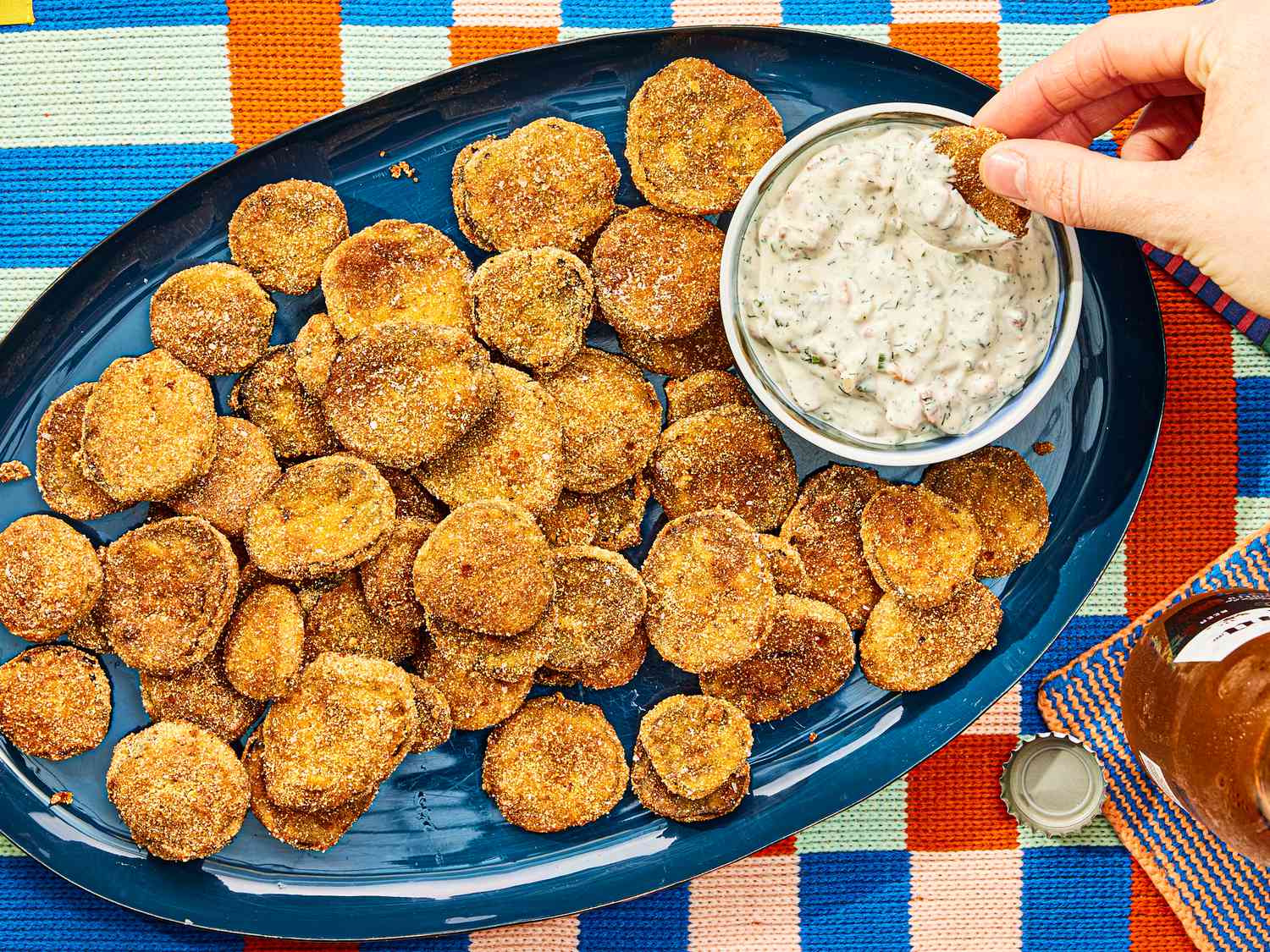 Blue platter with fried pickle and a small bowl of dip, with a hand dipping one pickle 