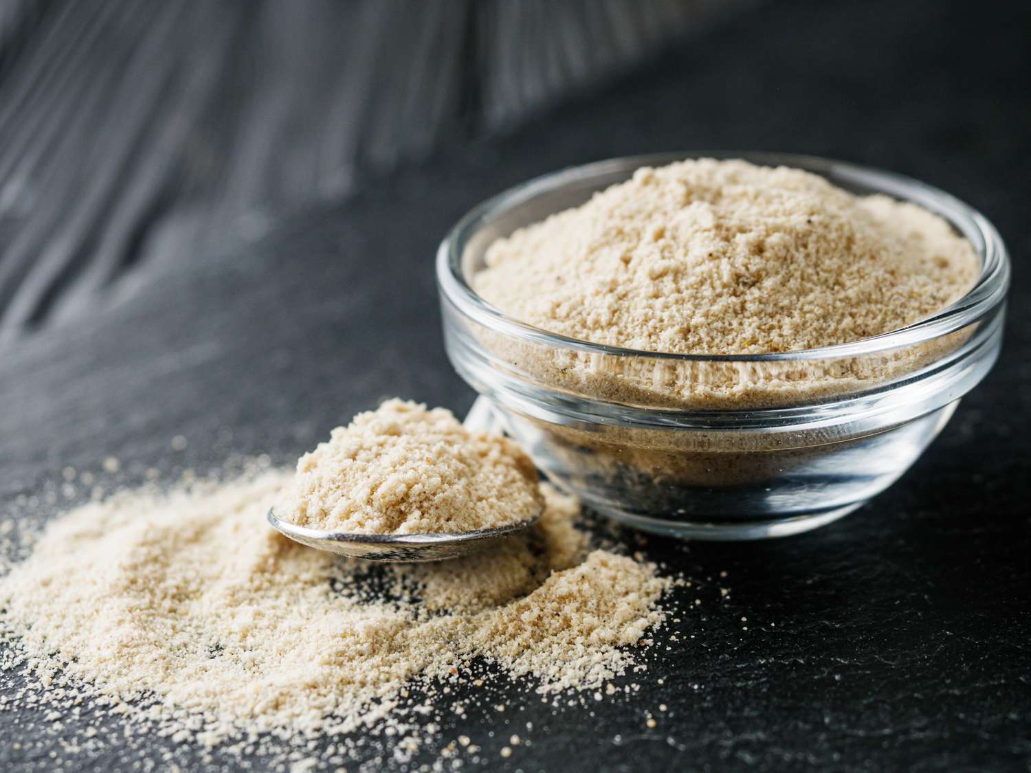 A glass bowl filled with a powdered ingredient with some of the powder spilled beside a metal spoon on a dark surface