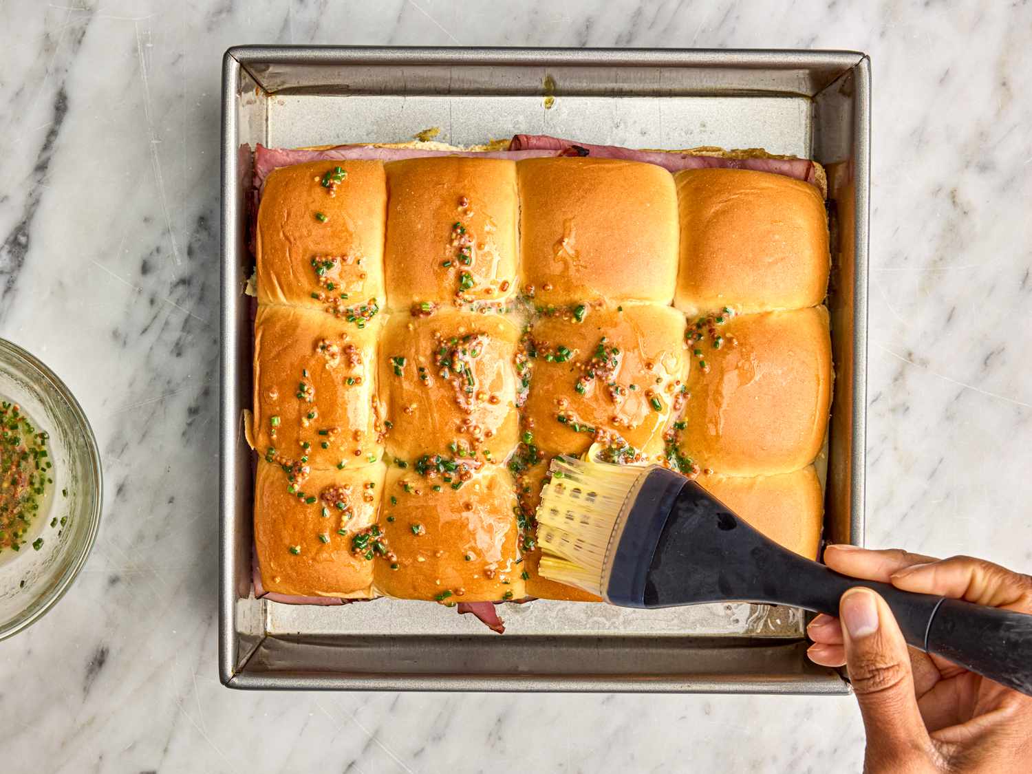 Baking dish with ham and cheese sliders being brushed with butter mixture