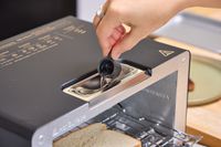 A hand pouring water into a slot in a balmuda toaster with a piece of bread visible inside