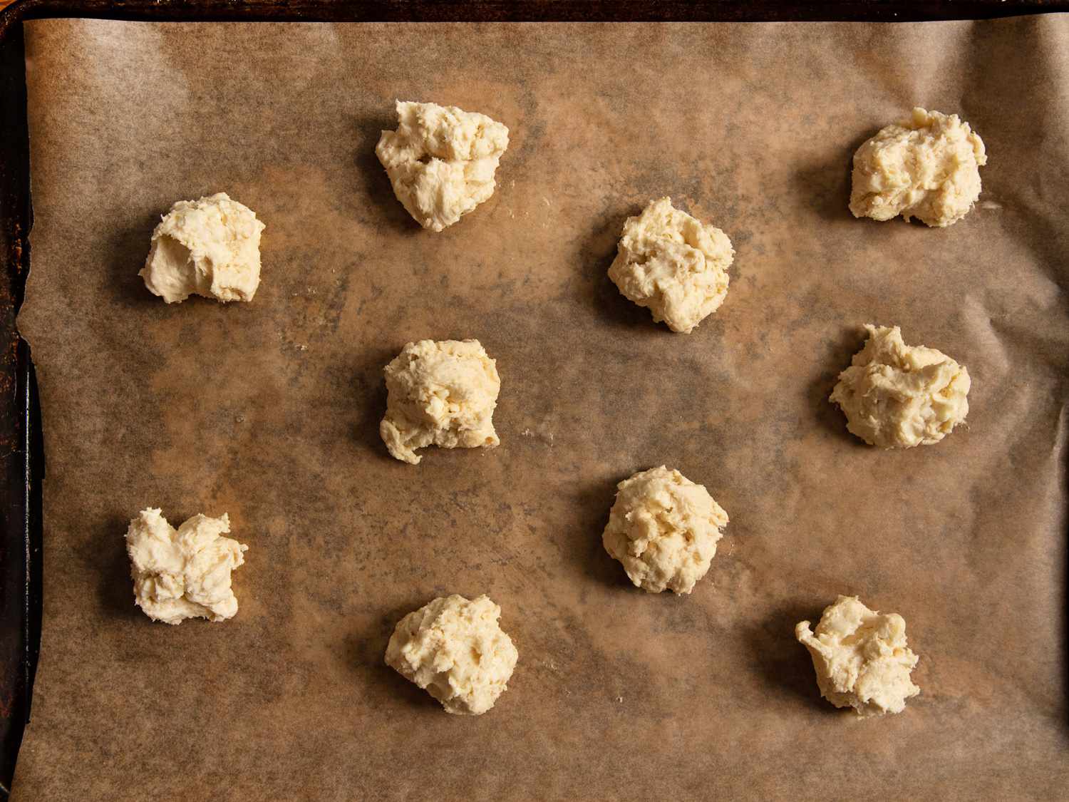 Dollops of biscuit dough on a parchment-lined rimmed baking sheet.