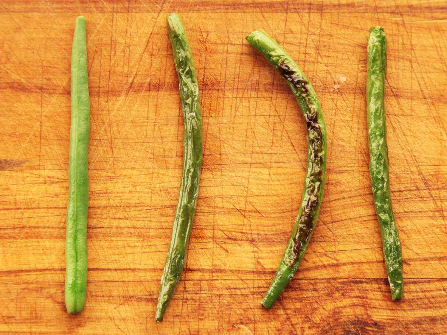 Overhead close-up of four green beans on a cutting board. Each has been cooked with a different method to simulate dry-frying.