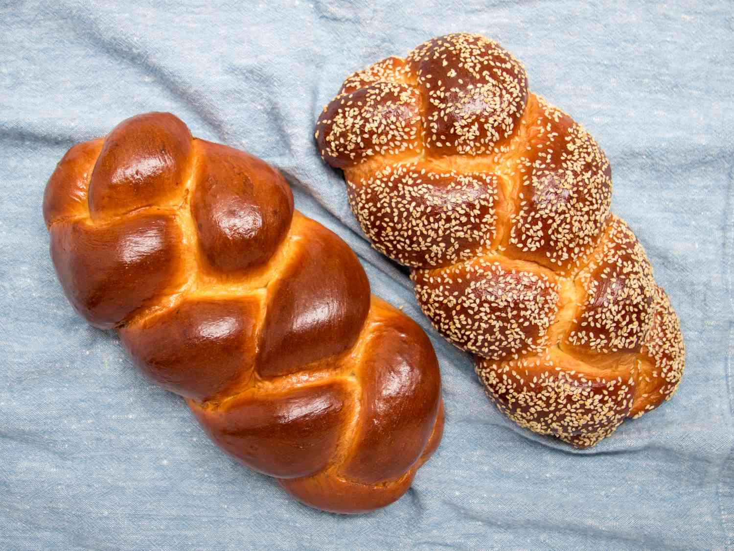 2 loaves of challah on a light blue textile. One challah has sesame seeds, the other is plain. 