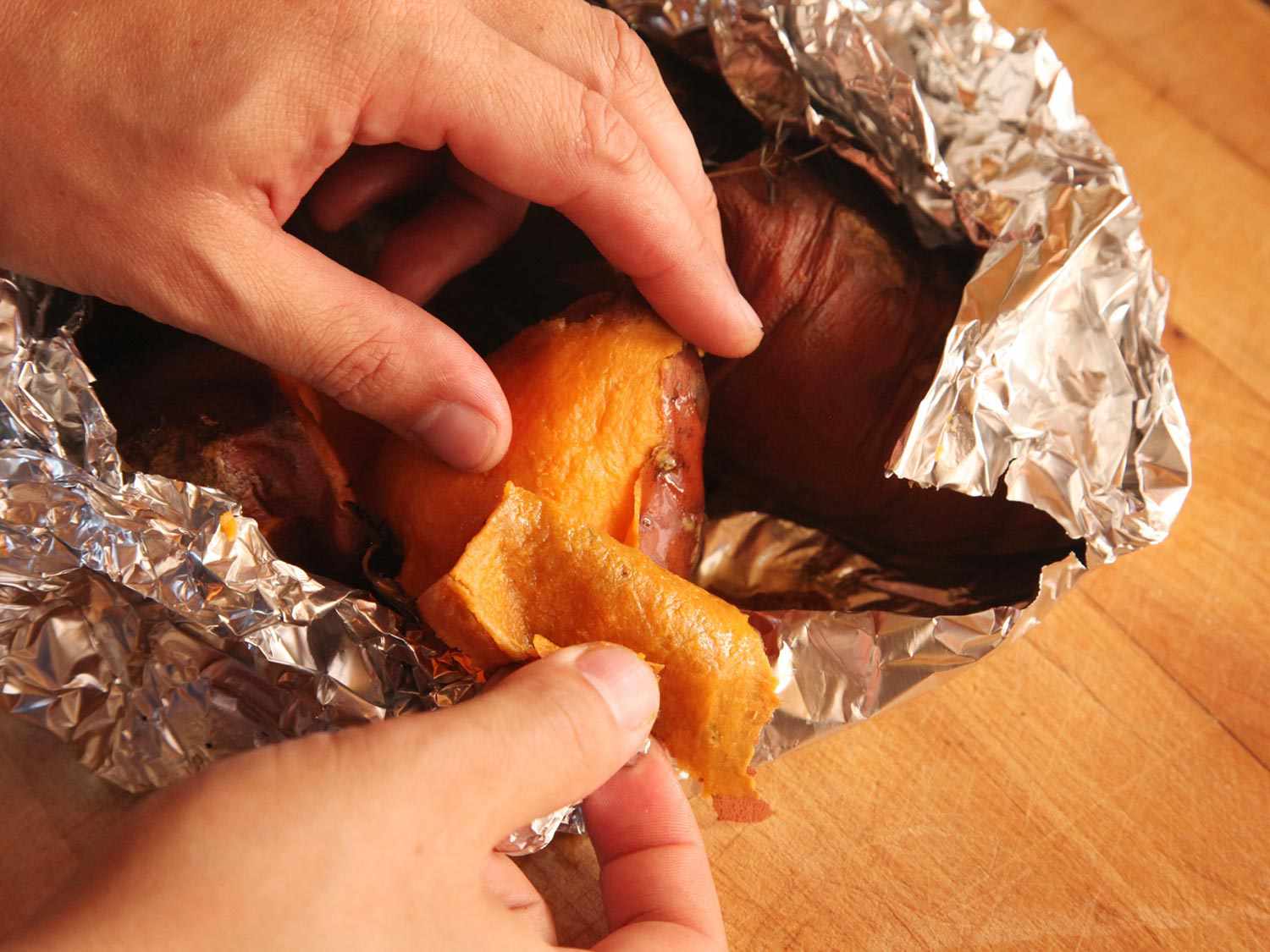 Hands peeling roasted sweet potatoes in tin foil