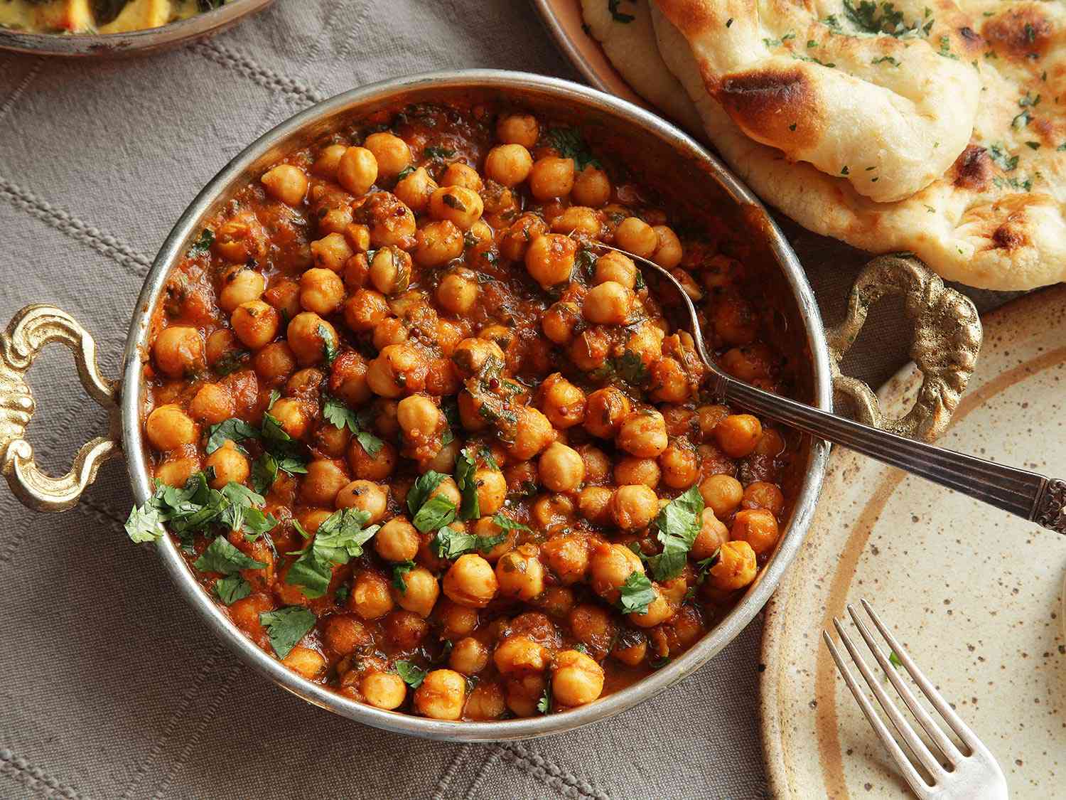 Cilantro-strewn channa masala in a shallow serving bowl with ornate brass handles, apparently Turkish in origin. A dish of freshly baked naan is close at hand.