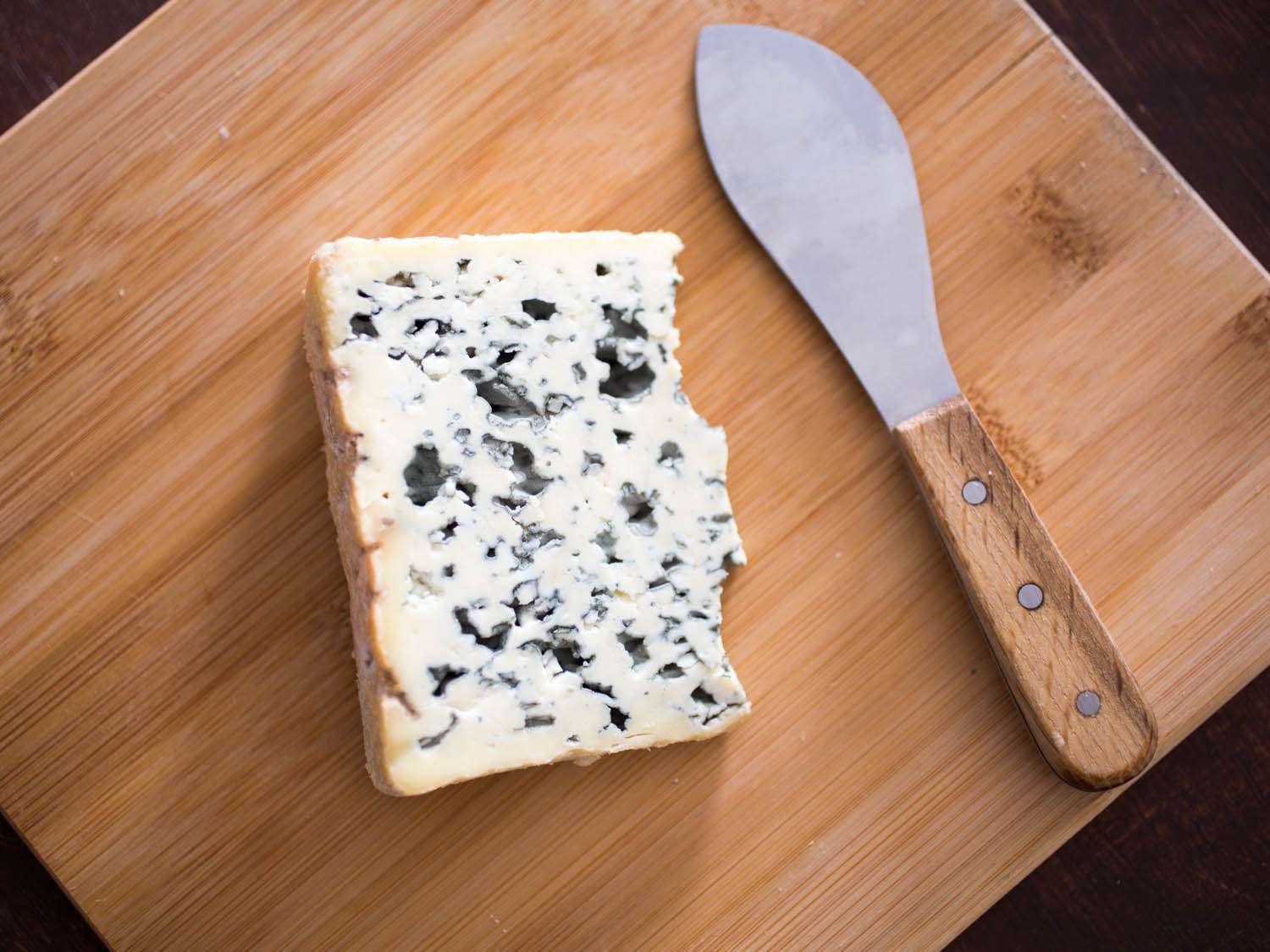 A thick wedge of Bleu d'Auvergne on a cutting board next to a cheese knife.