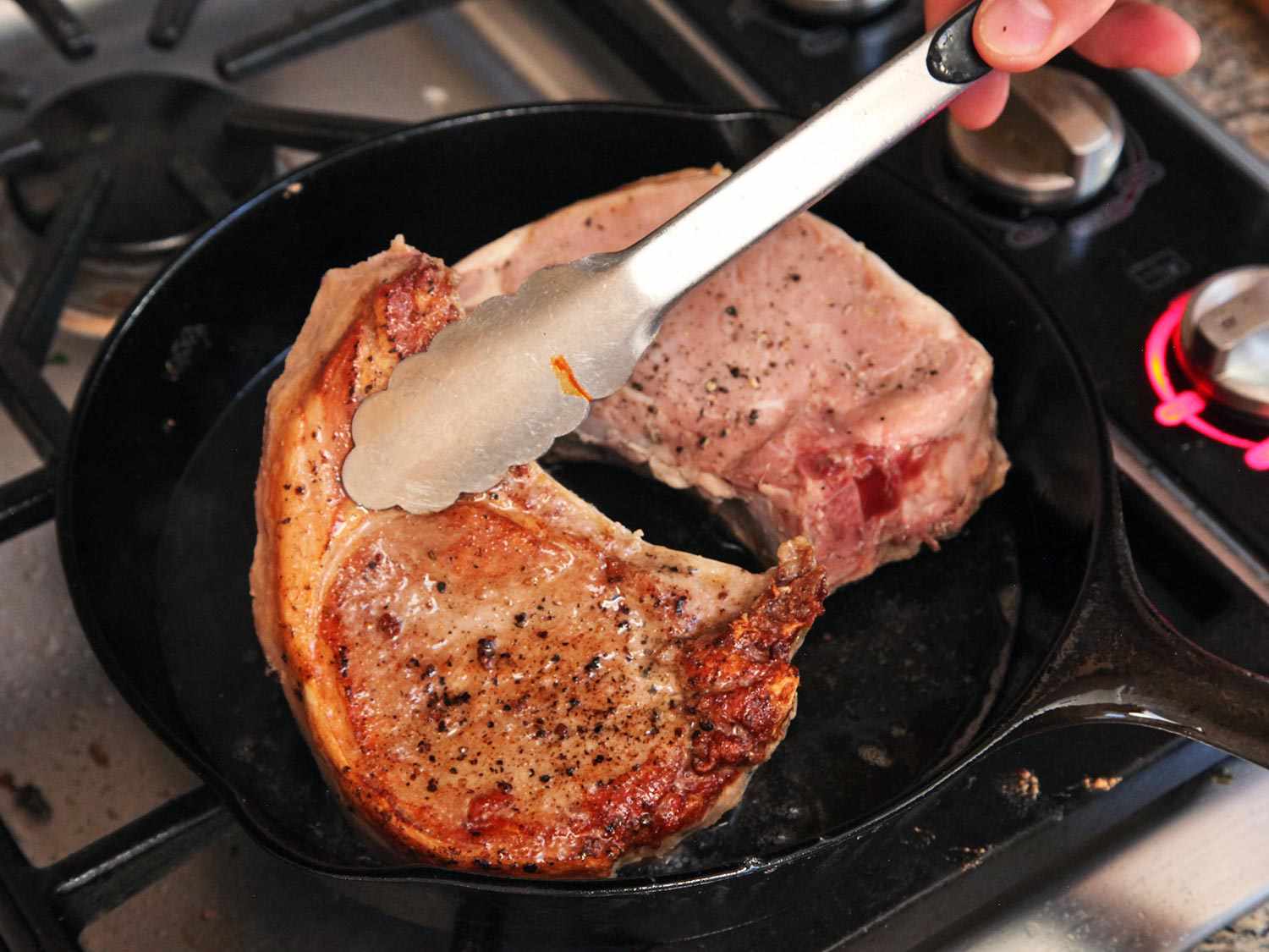 Two pork chops being cooked and flipped with tongs on a cast iron skillet. 