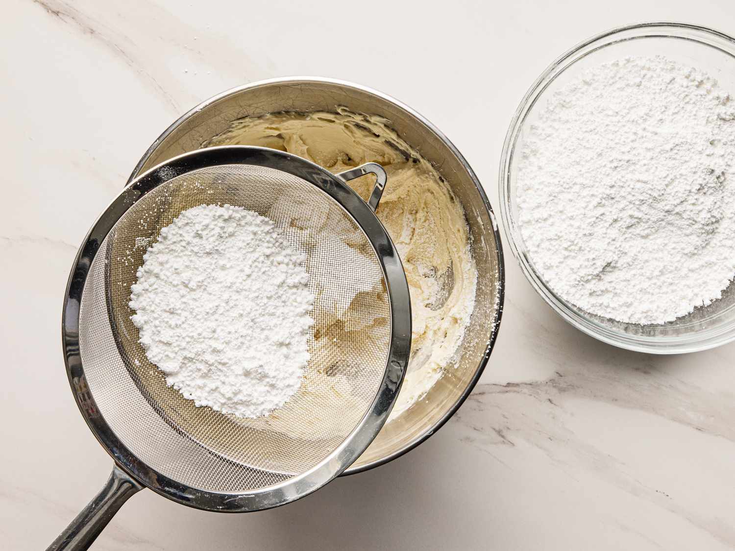 Sifting powdered sugar into a bowl of cake batter next to a bowl of powdered sugar