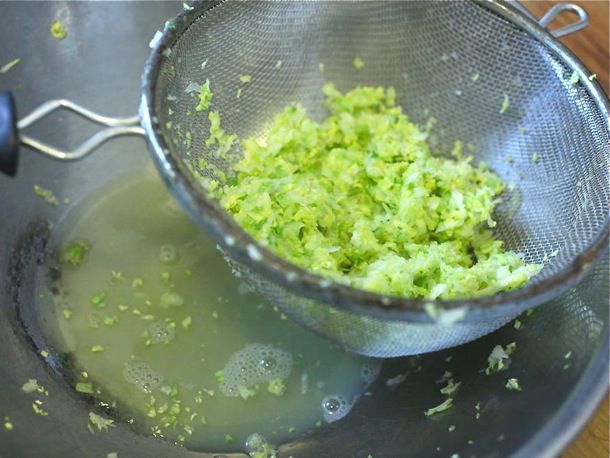 Chopped cabbage being purged of liquid in a mesh strainer above a large metal mixing bowl.