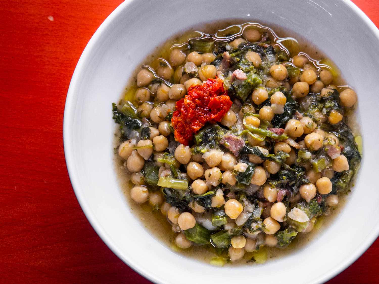 Overhead view of Chickpea Stew With Bacon and Broccoli Rabe, served in a white bowl on a red sureface.