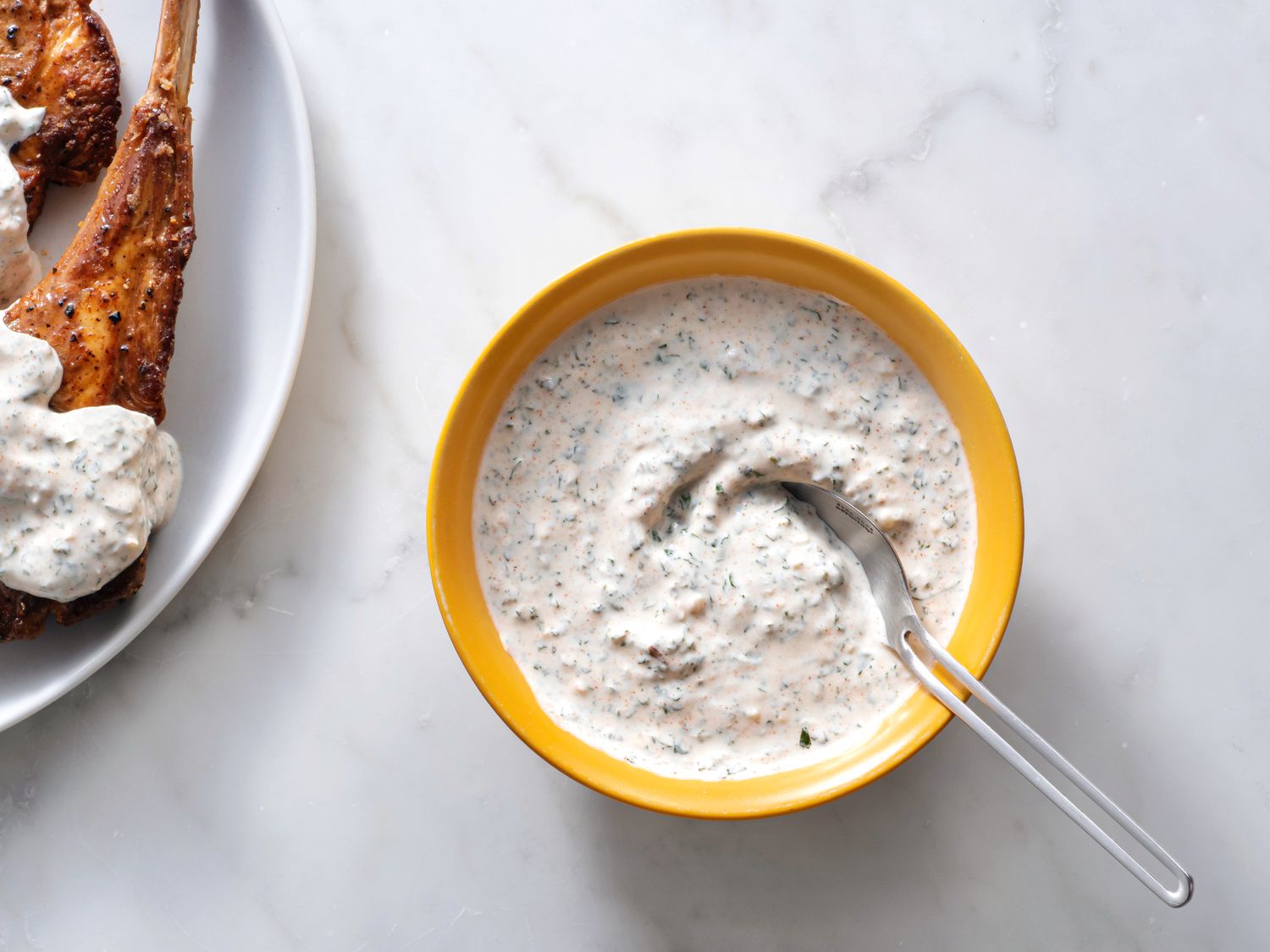 Yogurt mint sauce in a yellow bowl with a spoon, with sauce-covered meat on the left hand border of the image.