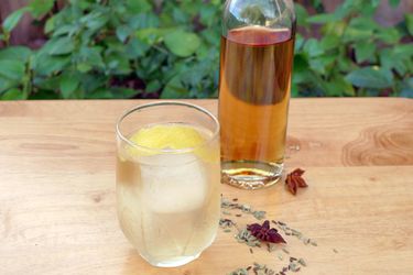 A stirred cocktail with as lemon twist sits next to a bottle of DIY aquavit. Some of the botanicals used in the recipe are scattered on the table nearby.