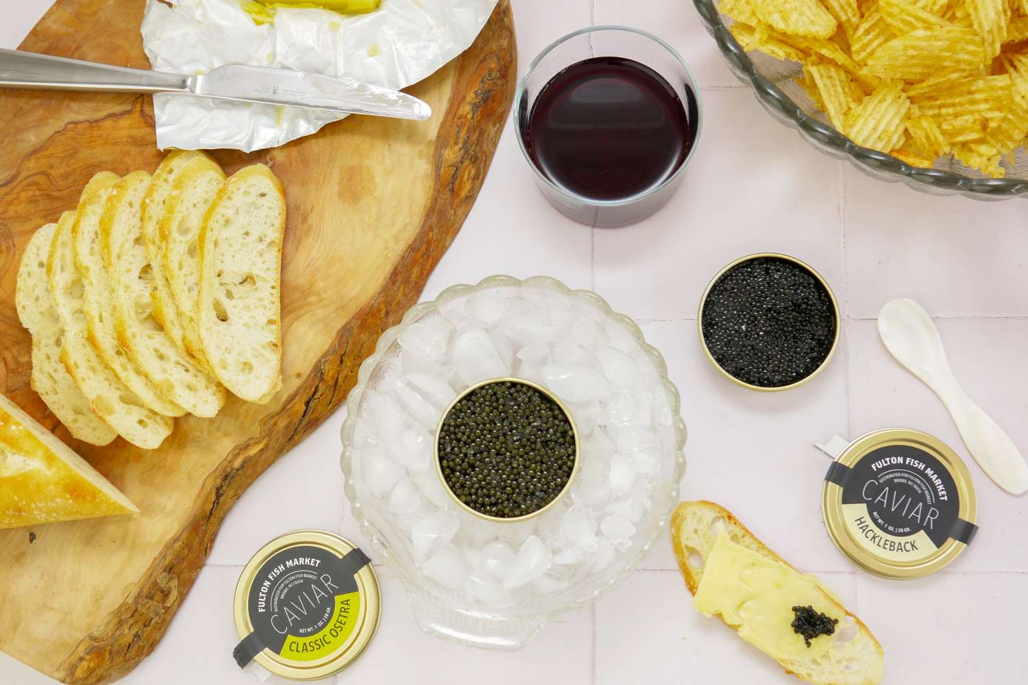 A spread on a table featuring bread slices caviar tins with a bowl of ice chips and a glass of wine