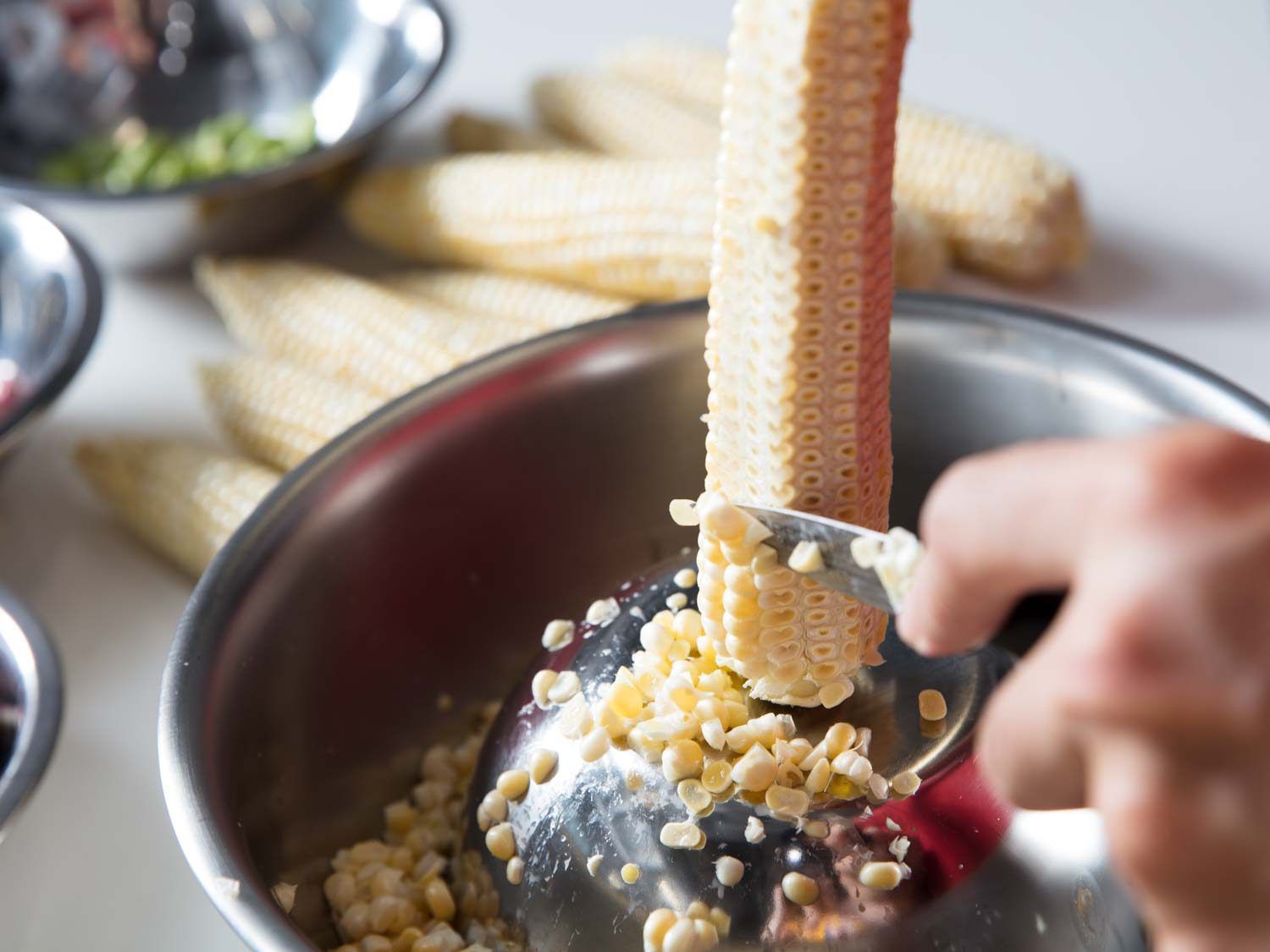 Cutting corn off the cob into a metal bowl, with the cob end resting on an upturned small metal bowl inside the larger one.