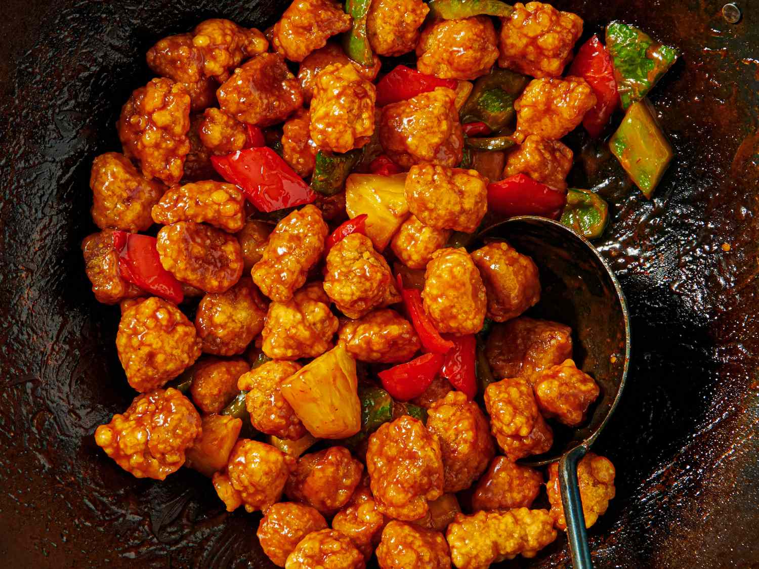 Sweet and sour pork served in a pan with a spoon includes vegetables like bell peppers and pineapple chunks