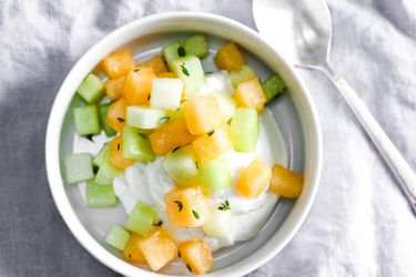Overhead view of a white bowl containing fresh cantaloupe and honeydew salad with lemon and thyme, served over a scoop of high-quality ricotta.