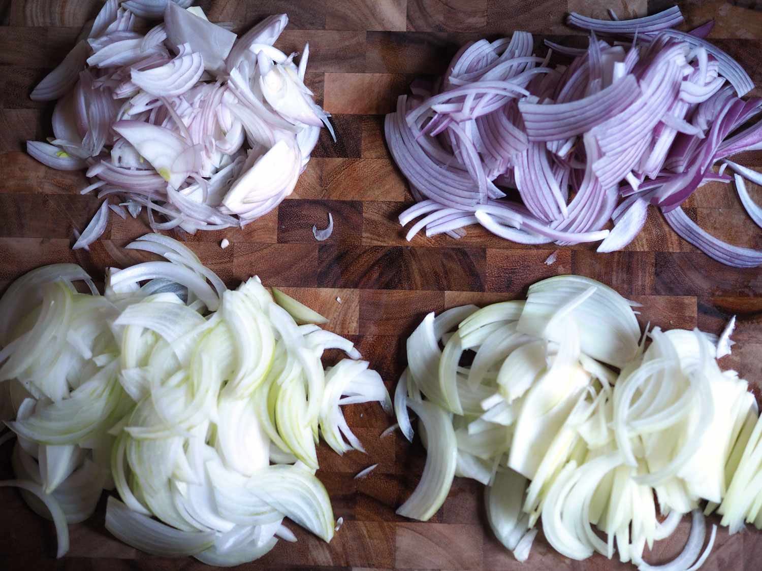 Sliced shallots, red onions, yellow onions, and sweet onions on a wooden cutting board.