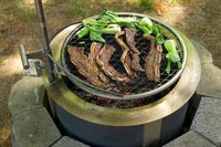 Meat and vegetables grilling over an open flame on a circular outdoor barbeque setup