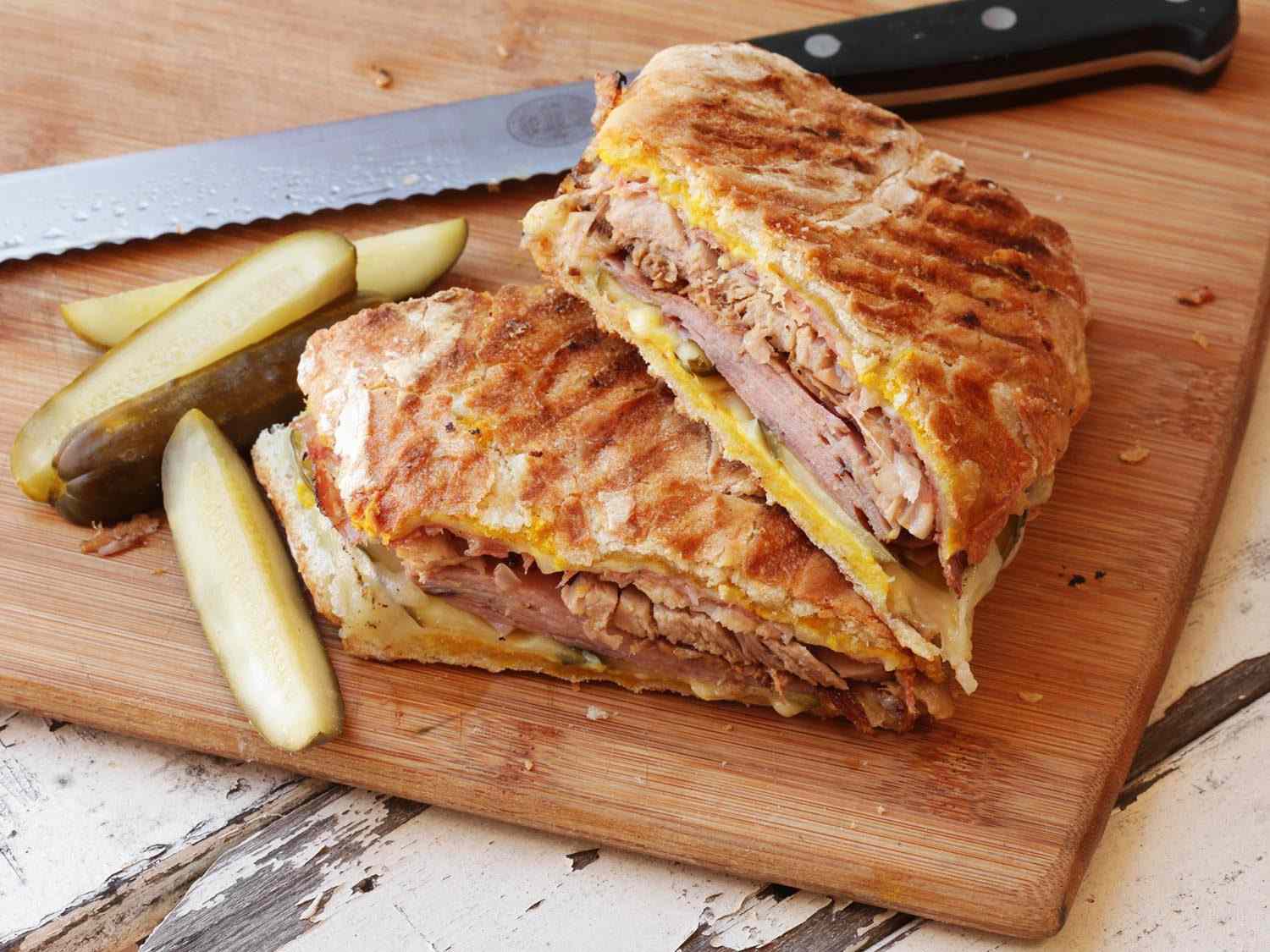 A grilled Cuban sandwich cut in half on a wooden cutting board with pickle spears.