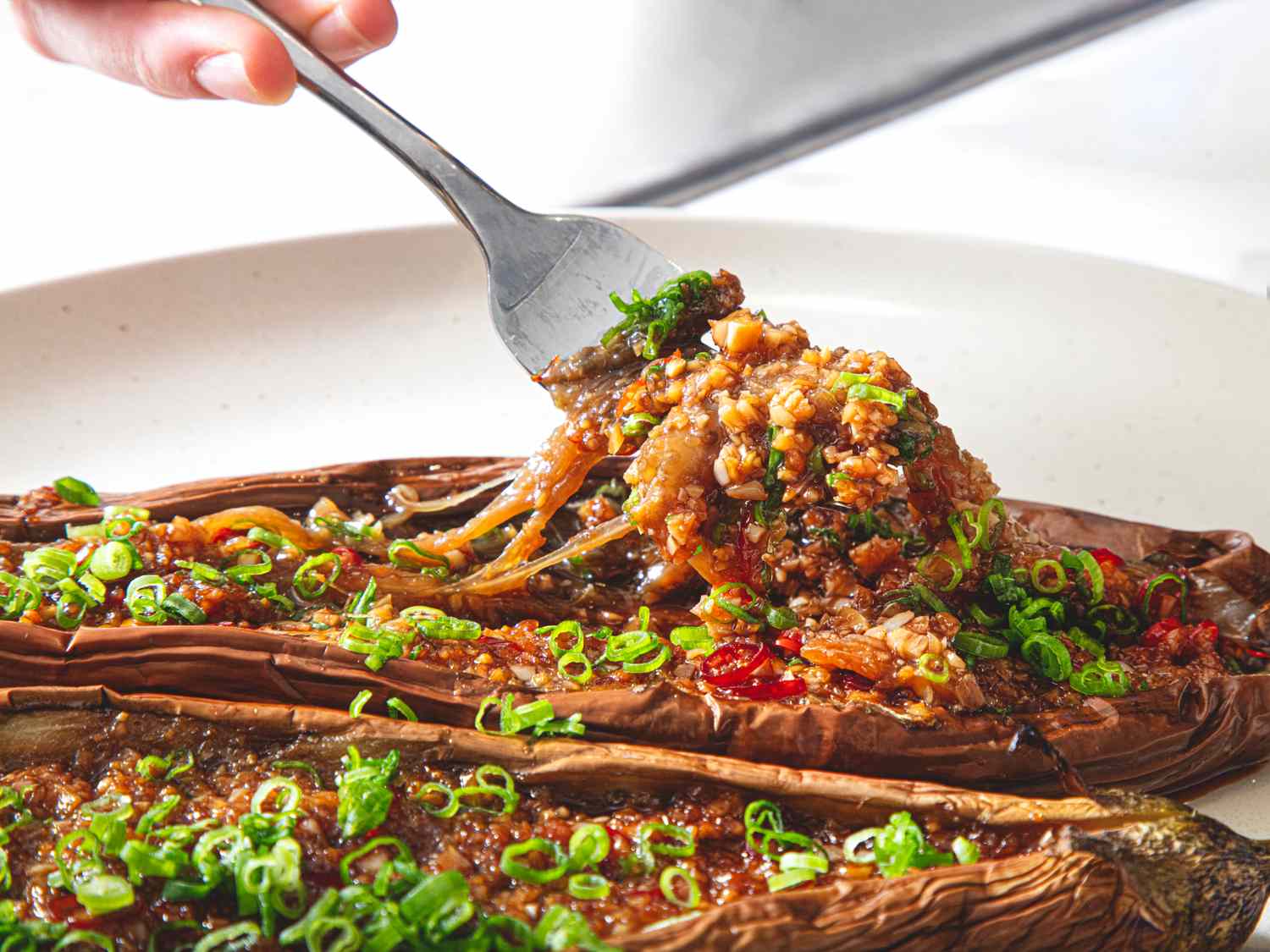 Roasted eggplants topped with savory sauce and garnished with scallions a hand using a fork to serve