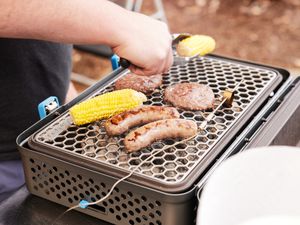 A person grilling burgers, corn, and sausages on a portable charcoal grill.
