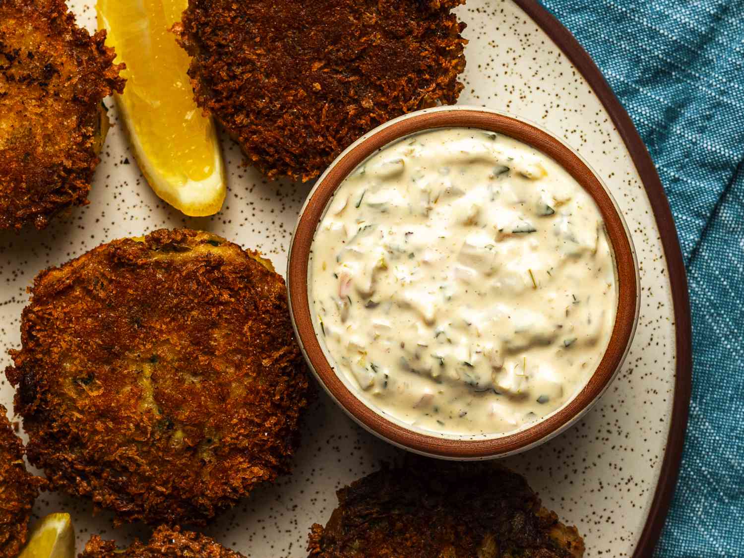 A small bowl of extra tangy tartar sauce on a speckled ceramic plate. The tartar sauce has crab cakes and lemon wedges around it.