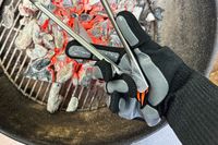 A person holds tongs over hot coals while wearing the Blackstone Griddle Gloves
