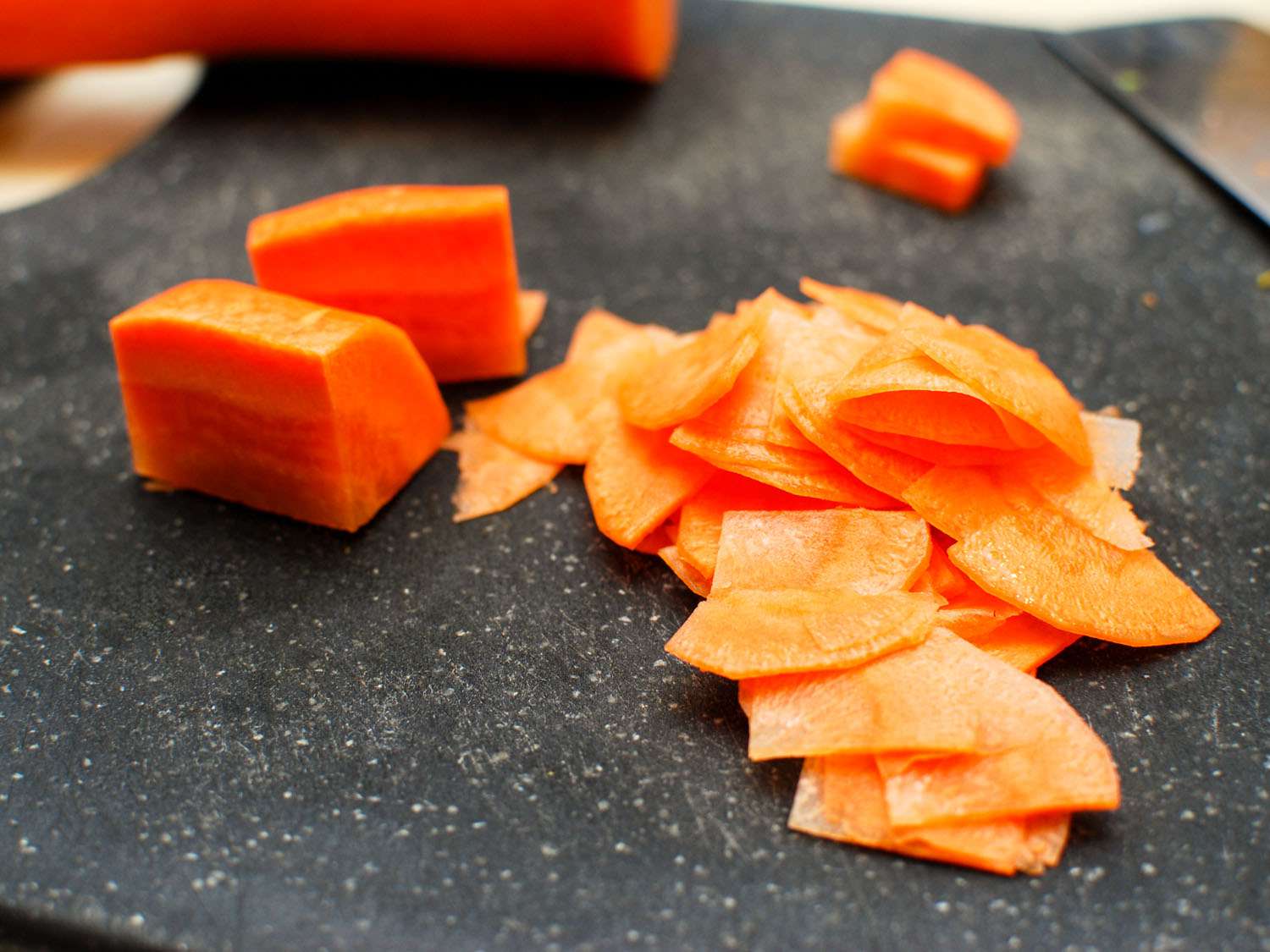 Several chunks of carrot that have been quartered lengthwise pictured on a cutting board, as well as a pile of thinly-sliced carrot quarter-moons.