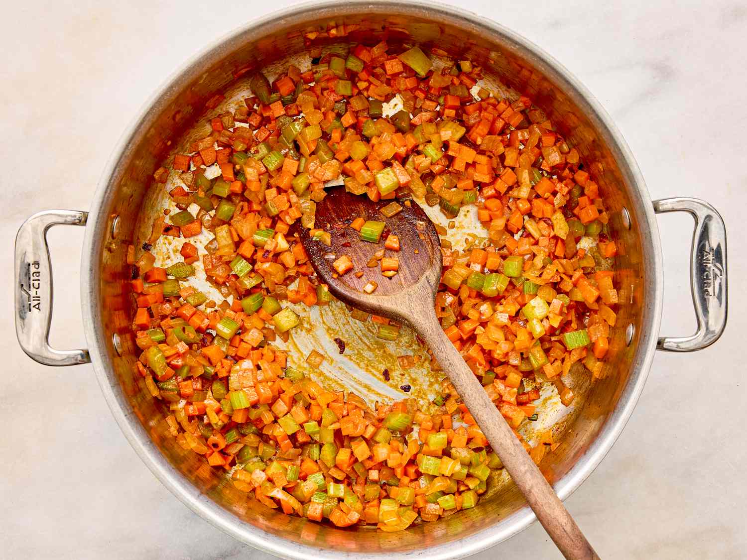 A pot on a marble surface containing diced vegetables being sauted with a wooden spoon