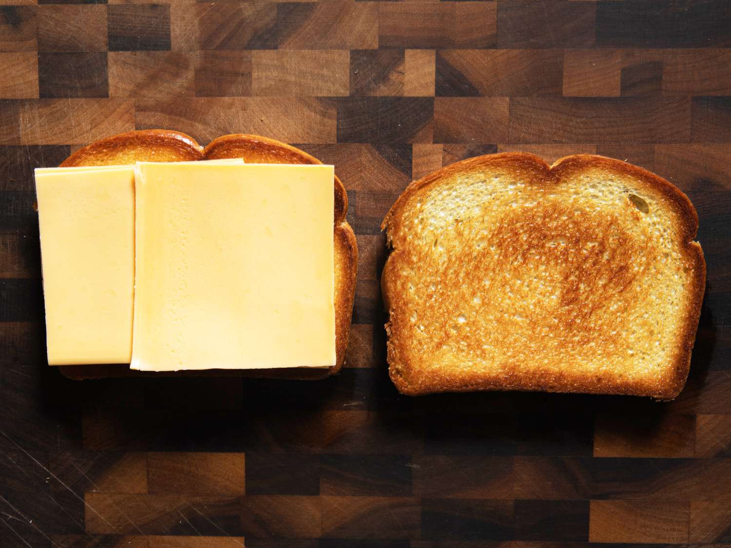 Two pieces of bread, tossed side up on a cutting board, with two pieces of yellow American cheese on one side