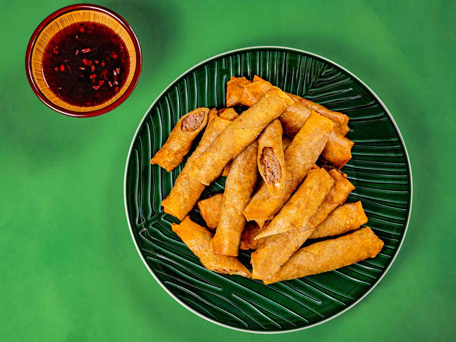 A green platter of lumpiang shanghai with dipping sauce on a green background.