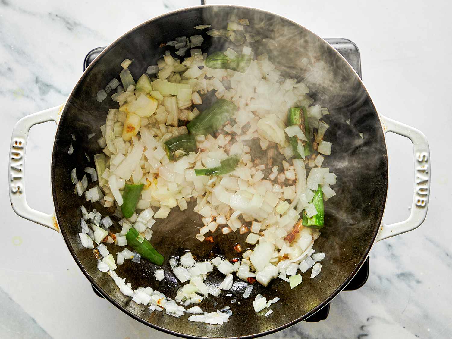 A pot on a stovetop containing chopped onions and green peppers being sauted