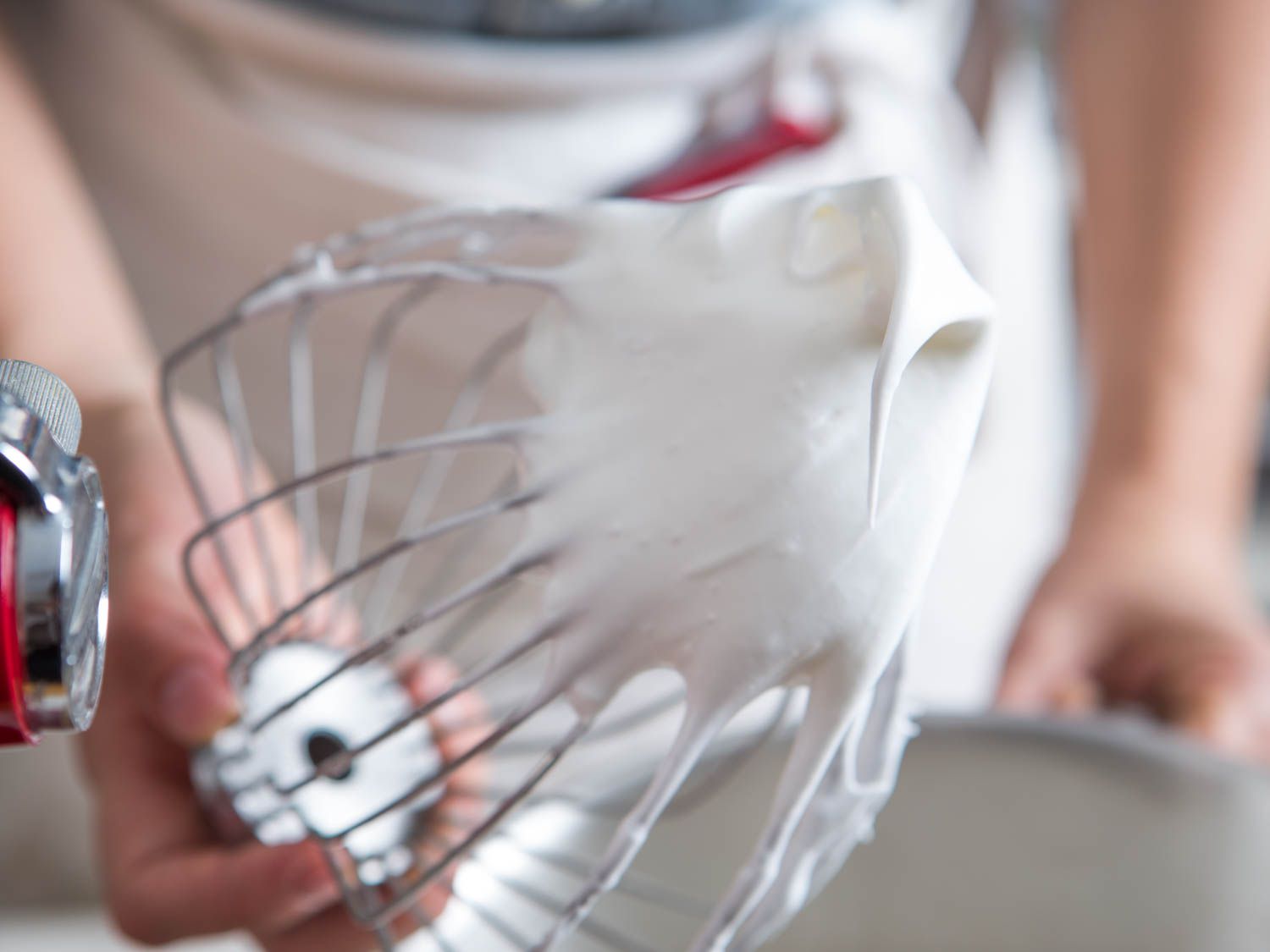 Hand holding a stand mixer whisk attachment with stiffly beaten egg whites and sugar on it.