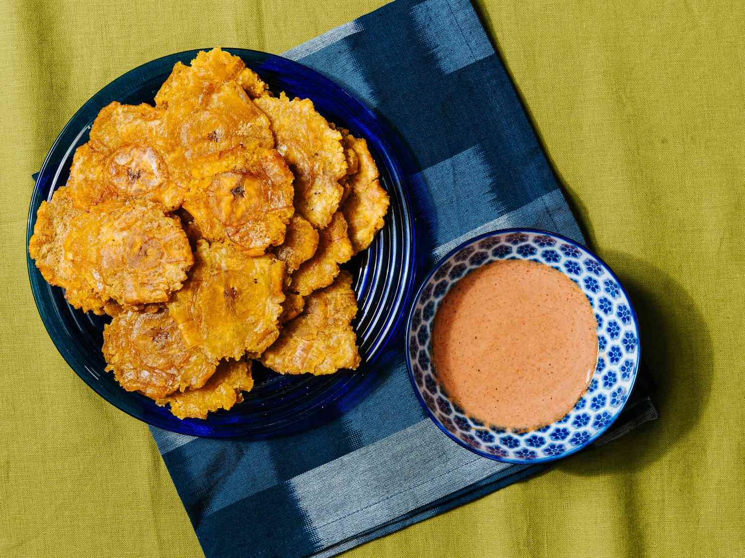 Overhead view of tostones on a blue plate with a blue napkin and bowl of dipping sauce