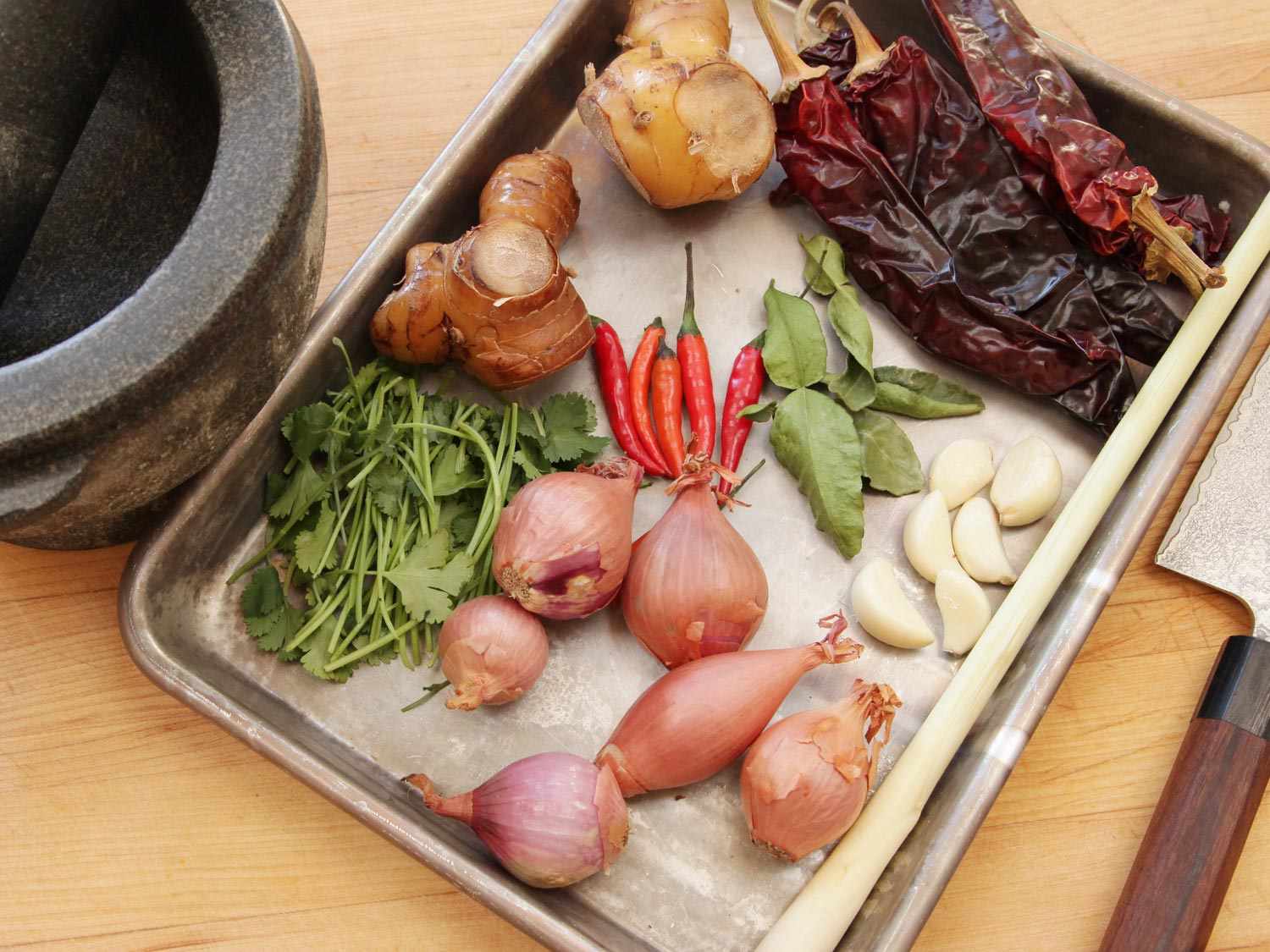 Aromatics assembled for phat phrik khing on a rimmed baking sheet: chiles, shallots, gallangal, makrut lime leaves, garlic. The baking sheet is next to a mortar and a knife. 