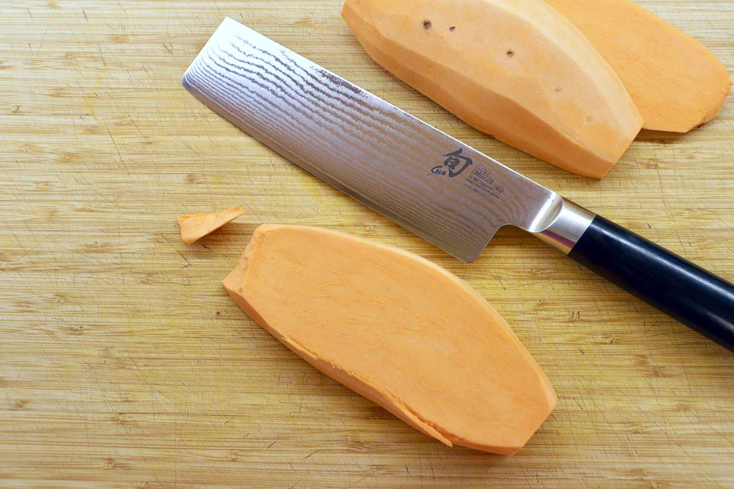 A damascus nakiri knife on a wooden cutting board with slices of sweet potato surrounding it