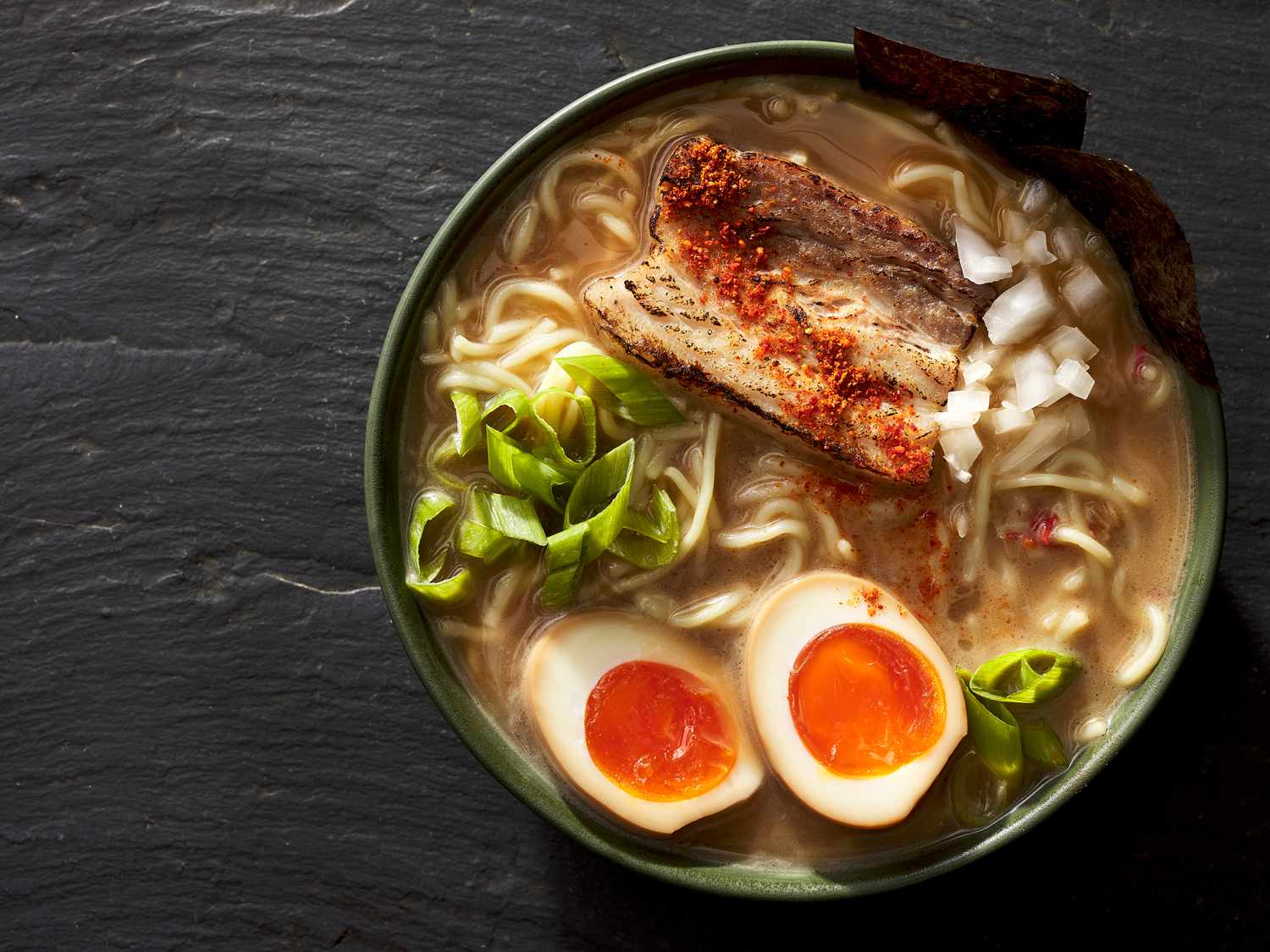 Bowl of ramen garnished with meat, eggs, and green onions on a black stone surface 