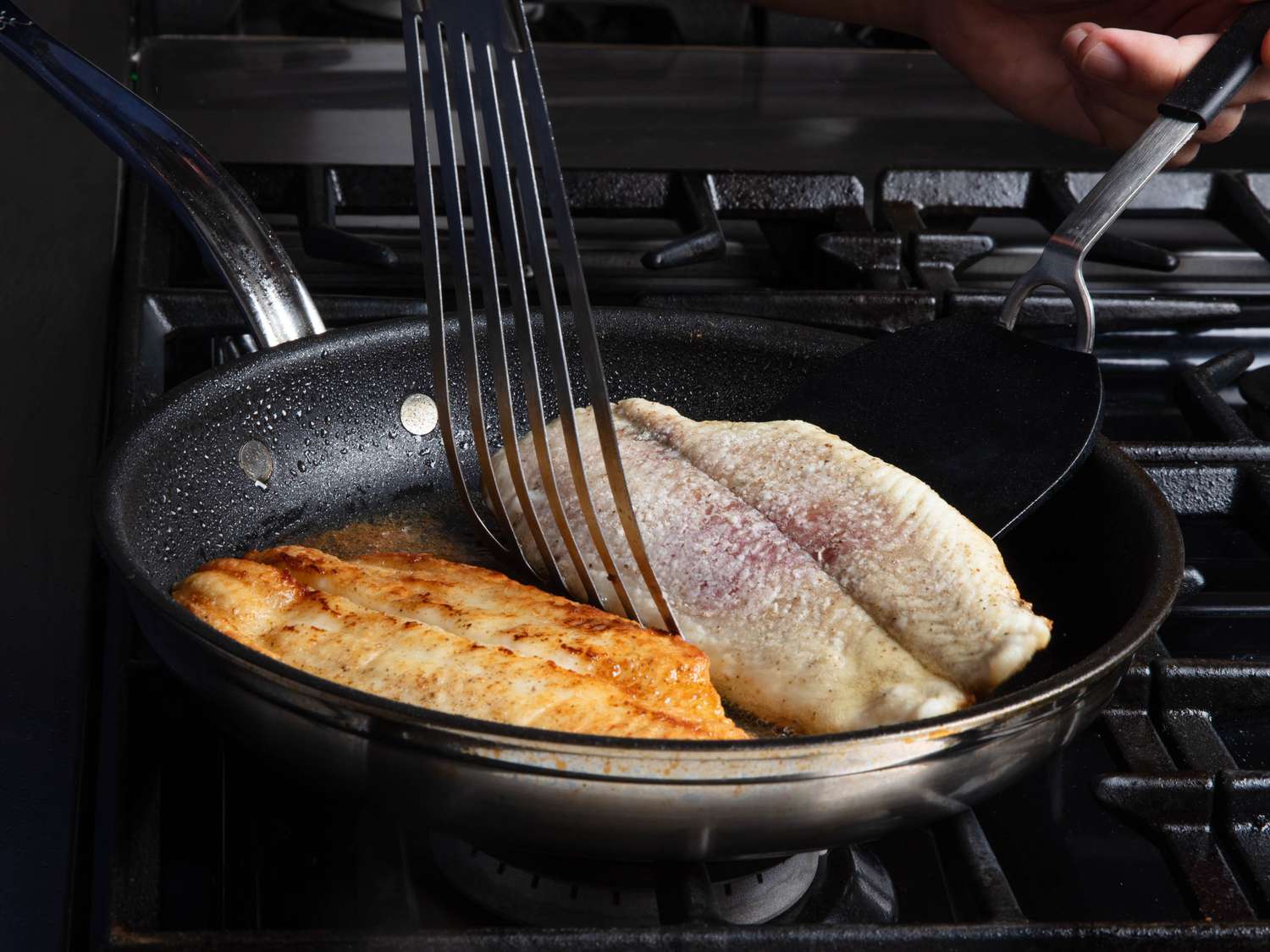 Two fish fillets sit in a nonstick skillet. One has been flipped over already and is a rich brown color; the other is in the process of being flipped using two large spatulas.