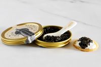 A tin of Marky's Beluga Caviar on a countertop, next to a prepared blini topped with caviar
